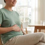 Une femme souriante assise dans son salon, la main posée sur son ventre, dans une atmosphère paisible.