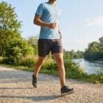 Un homme en tenue de sport court sur un chemin en terre au bord d'une rivière par une journée ensoleillée.